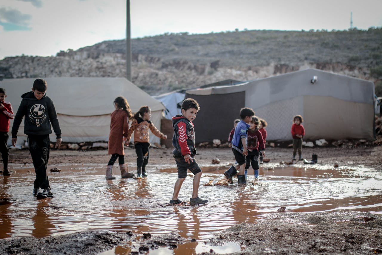 heros-img Kids in a refugee camp in Idlib, Syria, playing in muddy water, reflecting resilience and community spirit.