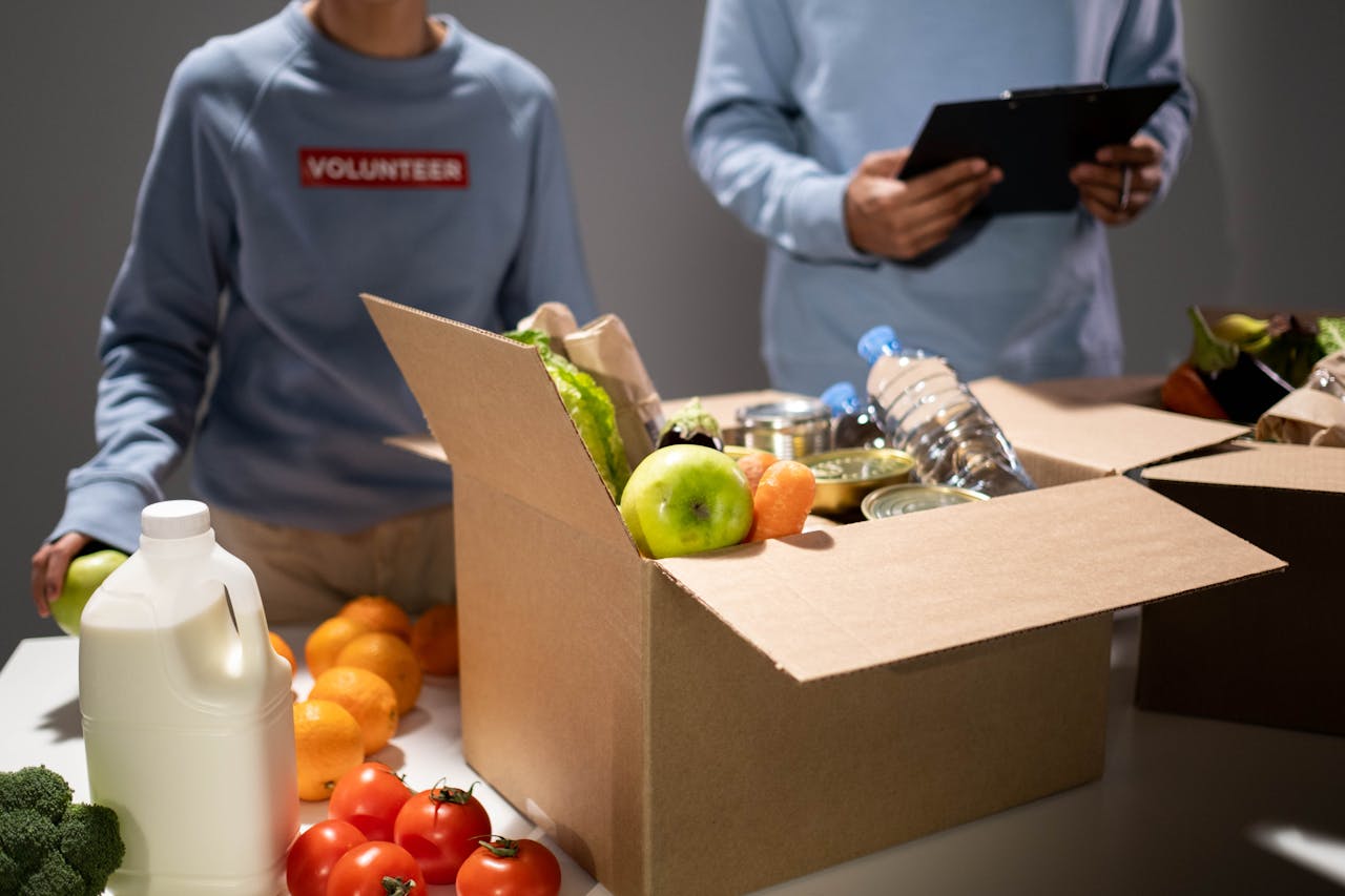 our-story People packing a cardboard box with essentials like fruits, vegetables, and bottled water for charity.
